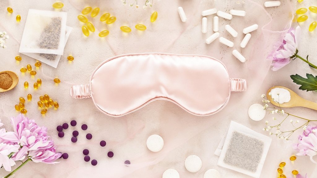 Tapa olho para dormir rodeado de diferentes tipos de cápsulas, inclusive cápsulas gelatinosas, além de duas colheres de pau com nutrientes em pó, flores e saquinhos de chá